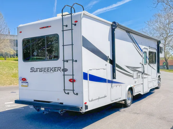 White Sunseeker RV, an ideal rv rental, parked outdoors. Rear view shows ladder, window. Side has a sleek black-and-blue design & awning.. Forest River Sunseeker 2021