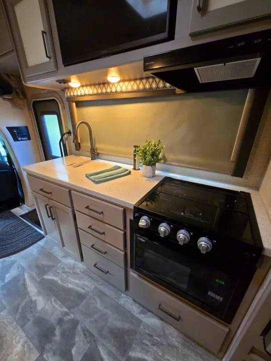 Inside an RV rental, a modern kitchen showcases light cabinets, a sink, black stove with oven, and faux-marble flooring.. Thor Quantum 2023