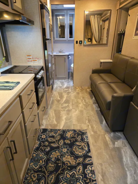 Modern RV rental: kitchen with light cabinets, sofa living area, and bathroom access. Features light floors and a dark rug.. Thor Quantum 2023