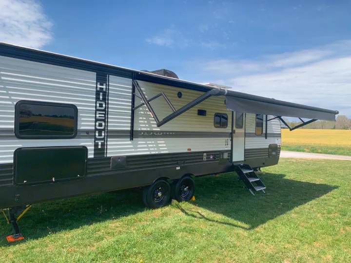 A modern travel trailer with an extended awning, ready for an RV rental adventure, parked on green grass under a blue sky.. Keystone Hideout 2022