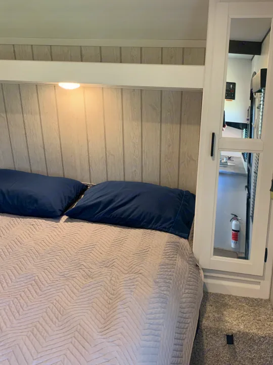 Cozy RV rental bedroom: bed with blue pillows, wood-paneled walls, and mirrored closet. A fire extinguisher is visible in reflection.. Keystone Hideout 2022