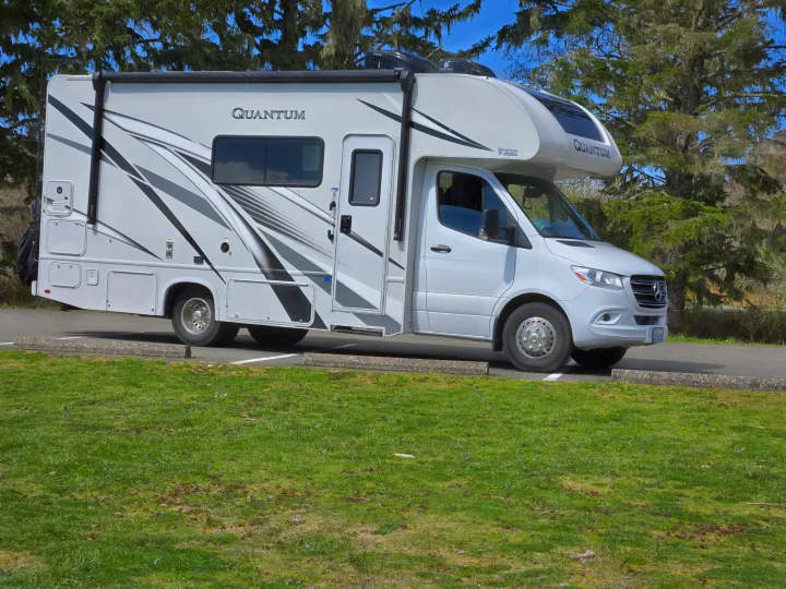 This white Quantum RV rental, a motorhome with gray accents, is parked on asphalt with green grass in the foreground and tall, mossy trees behind it.. Thor Quantum 2023