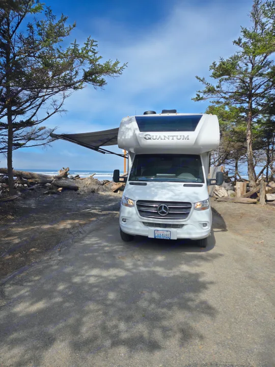 White Mercedes-Benz Quantum RV rental parked on a sandy path by an ocean beach with waves, an extended awning, and surrounding trees.. Thor Quantum 2023