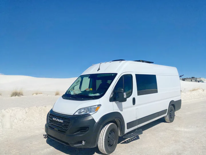 Bright white camper van RV rental parked on sandy ground with expansive white dunes and clear blue sky in the background.. RAM ProMaster 2023