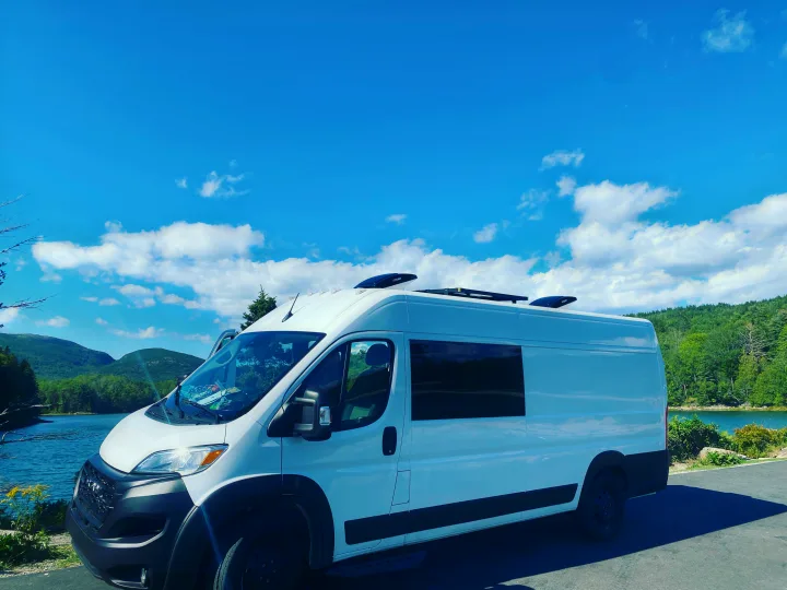Scenic RV rental adventure: White camper van parked by a tranquil lake with mountains under a clear blue sky.. RAM ProMaster 2023