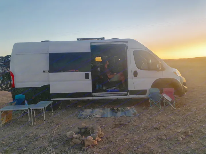 Relaxing at a desert RV rental campsite during sunset, with an open van door, a person inside, and outdoor seating around a fire pit.. RAM ProMaster 2023