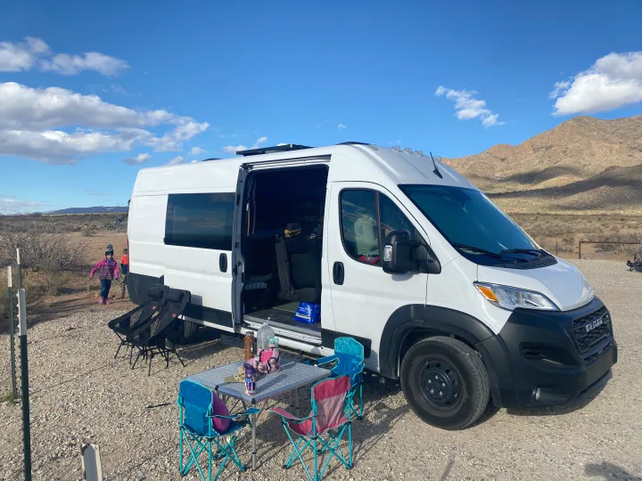 Family RV rental trip: A white camper van in a desert landscape, with an outdoor table and chairs setup.. RAM ProMaster 2023