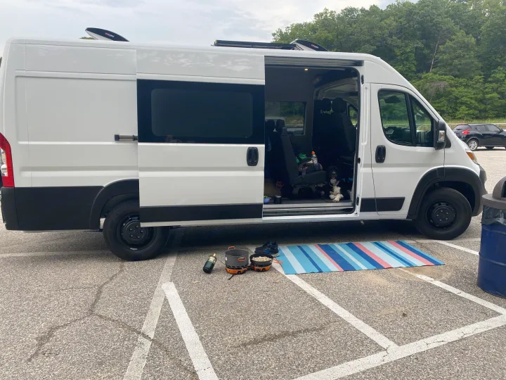 A white RV rental, a camper van, parked with its side door open, revealing a dog inside & outdoor cooking gear.. RAM ProMaster 2023