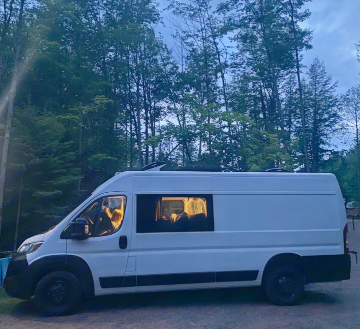 White camper van, perfect for RV rental, parked in a forest at dusk. Interior lights are on, showing people inside for cozy camping.. RAM ProMaster 2023