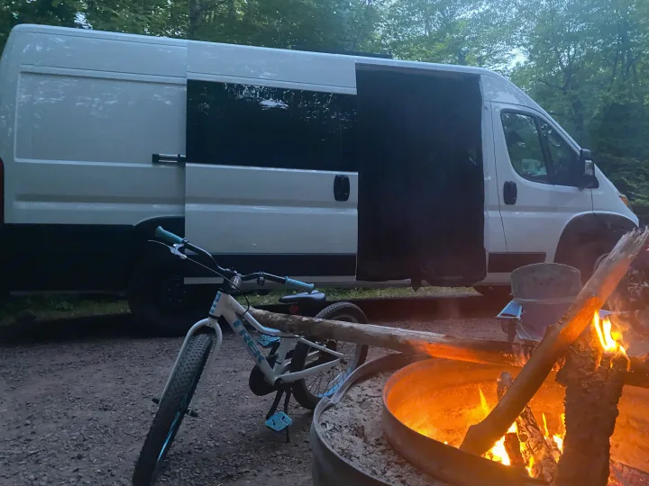 An RV rental camper van is parked at a dusky campsite, with a bicycle and a cozy campfire blazing in a metal pit.. RAM ProMaster 2023