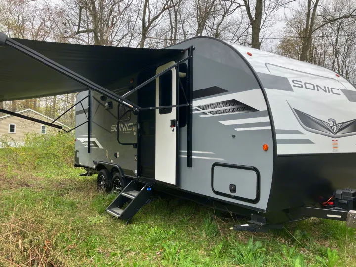 Modern gray and white Sonic RV rental with extended awning and open door, parked in a grassy area with trees.. Sonic Sonic Trailer 2023