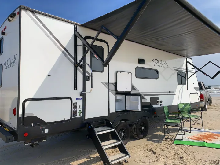 Spacious RV rental set up on a sandy beach, with awning, chairs, and an outdoor fridge, perfect for beach camping.. Dutchmen Kodiak Ultra Lite 2022