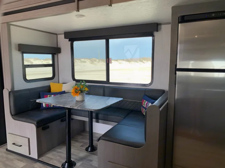 Inside an RV rental: a spacious dinette with black leatherette seating, a table, and windows overlooking sandy dunes and a fridge.. Dutchmen Kodiak Ultra Lite 2022