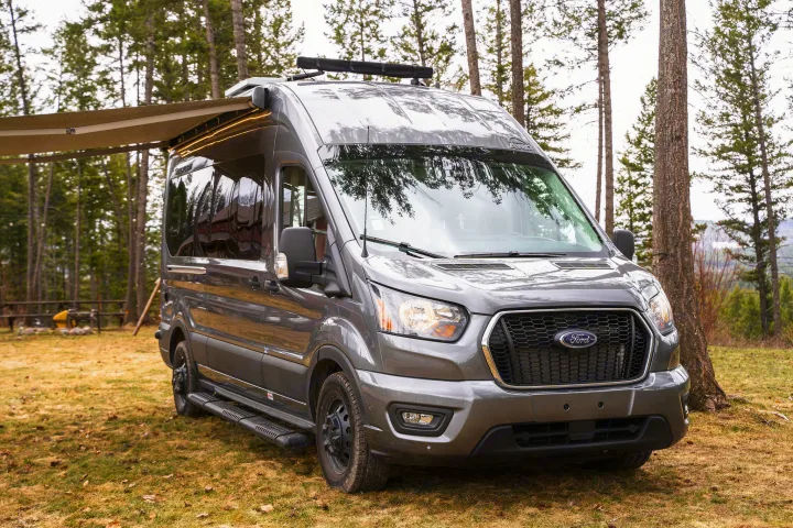 Grey Ford Transit camper van for rv rental, parked in a forest campsite with an extended awning & lights.. Thor Sanctuary 2023