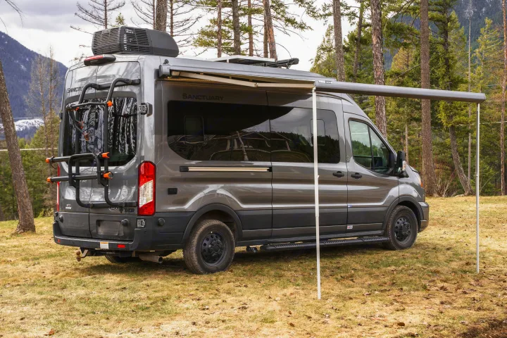 Modern gray rv rental van, "Sanctuary" model, with awning extended and bike rack, nestled in a scenic mountain forest.. Thor Sanctuary 2023