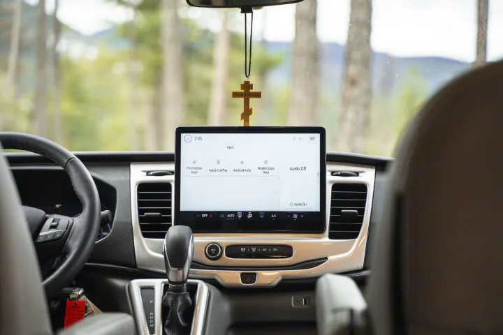 rv rental interior: Dashboard with a large central touchscreen, steering wheel, and gear shifter. A cross hangs, trees are visible outside.. Thor Sanctuary 2023
