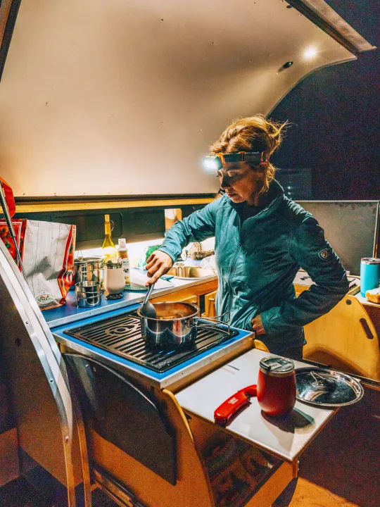 Inside an RV rental, a woman with a headlamp cooks dinner on a portable stove in her compact kitchen.. Pindrop Travel Trailer Fina Roma 2022
