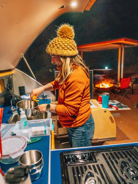 A woman washes dishes at the compact outdoor kitchen of an RV rental, a cozy campfire glowing in the background under the night sky.. Pindrop Travel Trailer Fina Roma 2022