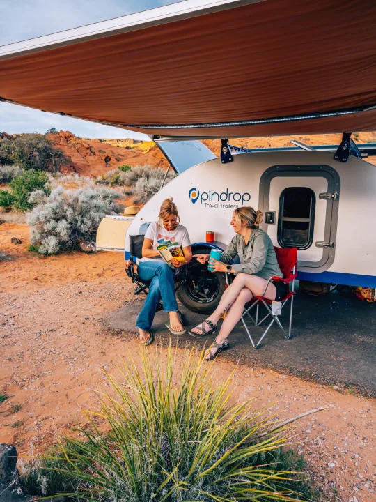 Two women relax under an awning next to a Pindrop rv rental travel trailer in the desert. One reads, the other sips a drink.. Pindrop Travel Trailer Fina Roma 2022