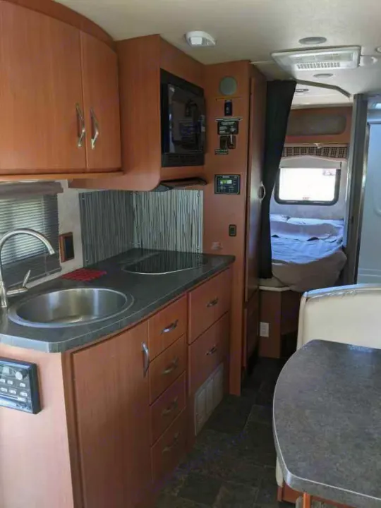 This RV rental interior features a kitchen with sink, cooktop, and microwave, leading to a private bedroom and dinette.. Itasca Navion 2012
