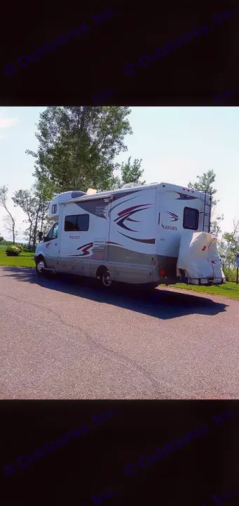 Explore the outdoors! This white Winnebago Navion RV rental is parked on asphalt with trees and water in the background.. Itasca Navion 2012