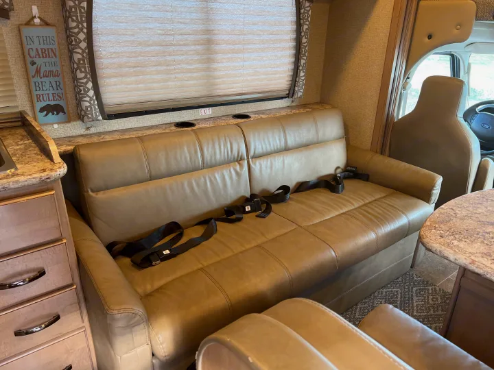 RV rental interior: A spacious tan sofa with seatbelts, an overhead window, and a decorative "Mama Bear Rules" sign.. Thor Chateau 2019