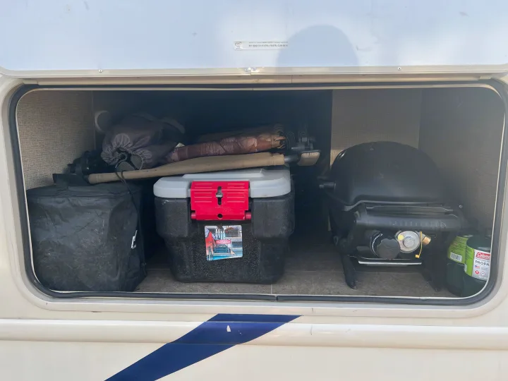 Inside an RV rental's exterior compartment: a camping grill, propane tanks, and a toolbox are neatly stored for travel.. Thor Chateau 2019
