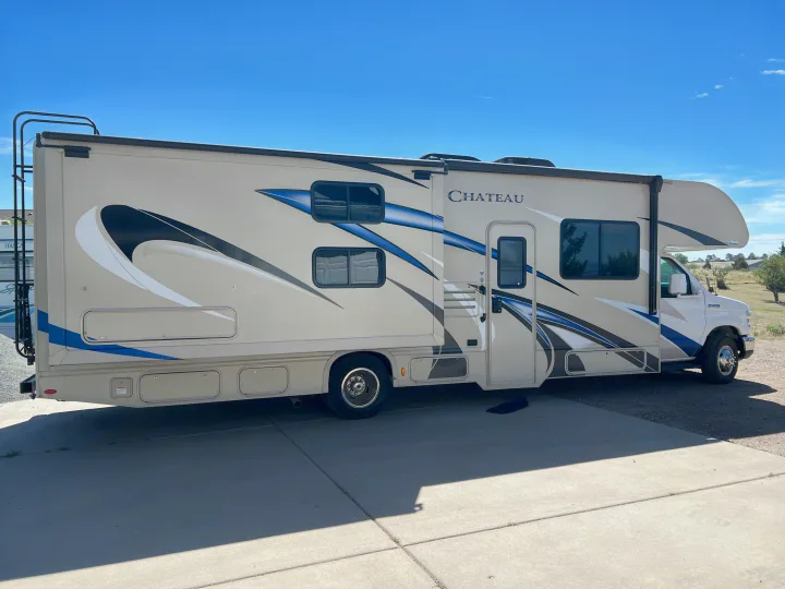 Side view of a beige Chateau RV rental, adorned with blue and black graphics, parked on concrete under a clear blue sky.. Thor Chateau 2019