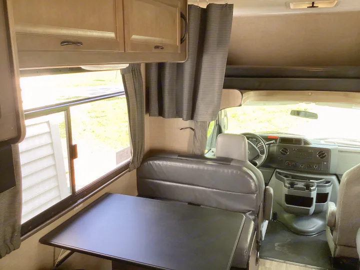 Interior of an RV rental, showing the driver's cab, dashboard, and the living area with a black table and bench seats.. Thor Majestic 2019