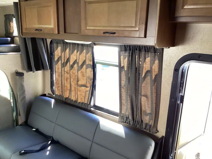 Bright RV rental interior featuring light wood cabinets, gray seating with seatbelts, and window curtains.. Thor Majestic 2019