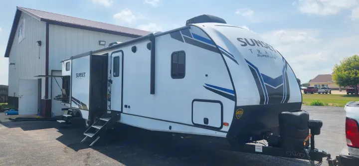 White Sunset Trail RV rental with an outdoor kitchen, steps extended, parked next to a building on a sunny day.. Crossroads Sunset Trail 2022