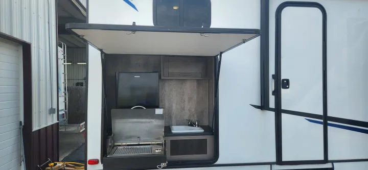 An RV rental showcasing its outdoor kitchen: a mini-fridge, grill, and sink are revealed behind an open hatch.. Crossroads Sunset Trail 2022