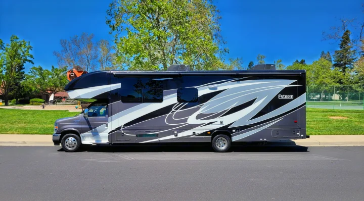 A sleek, dark gray and white Esteem Class C motorhome for rv rental, parked on a sunny day near a park and tennis courts.. Entegra Coach Esteem 2021
