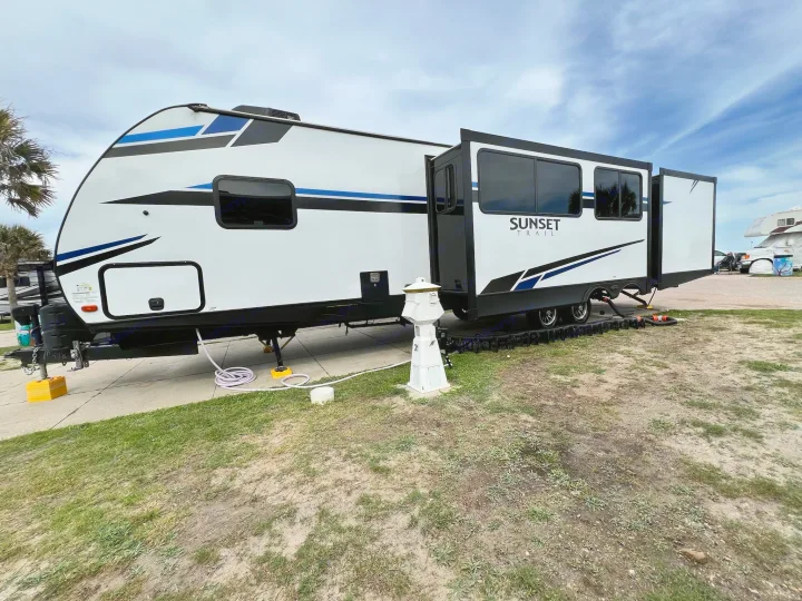 A modern white and blue Sunset Trail RV rental is fully set up at a spacious campsite, its slide-out extended.. Crossroads Sunset Trail 2021
