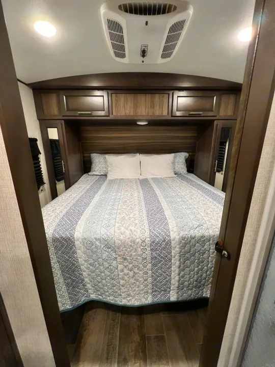 This RV rental bedroom features a comfy bed with a blue/white quilt, wood cabinets, and an overhead AC unit.. Crossroads Sunset Trail 2021
