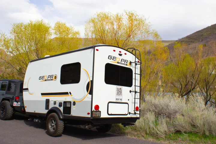 A white Geo Pro RV rental trailer hitched to a dark grey Jeep Wrangler, parked roadside amidst vibrant spring foliage.. Forest River Rockwood Geo Pro 2022