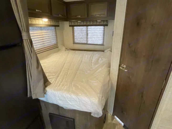 Inside an RV rental, showing a bed with a plastic-covered mattress, overhead cabinets, and windows with blinds.. Gulf Stream Conquest 2023