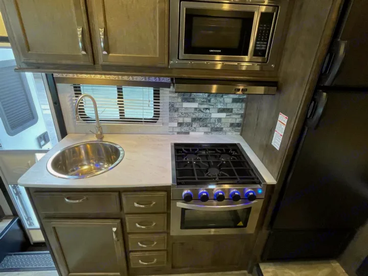 An inviting RV rental kitchen showcasing a stainless steel sink, 3-burner stove, oven, microwave, and light wood cabinets.. Gulf Stream Conquest 2023