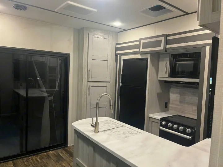 Spacious RV rental kitchen with modern light wood cabinets, an island sink, stove, microwave, & fridge. Sliding door access.. Keystone RV Fuzion Impact Toy Hauler 2022