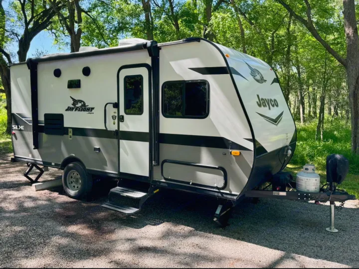 A white & grey Jayco Jay Flight SLX travel trailer, ideal for an RV rental, is set up on a gravel campsite amidst green trees.. Jayco Jay Flight SLX 2022