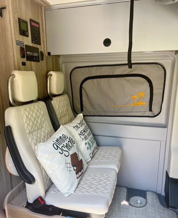 View of a modern RV rental interior featuring quilted seats, camping pillows, a control panel, and a palm tree window shade.. Winnebago Solis 2024