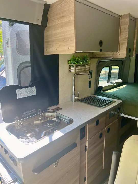 RV rental kitchen: This image shows a compact two-burner stove, sink, patterned countertop, and overhead storage in a camper van.. Winnebago Solis 2024
