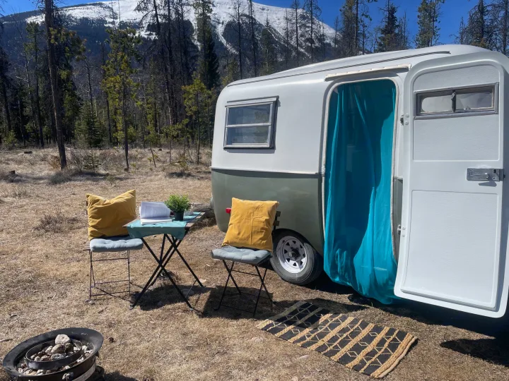 Cozy RV rental campsite in a mountain forest. Features a small camper, outdoor seating, and fire pit.. Boler Holitrai 1976