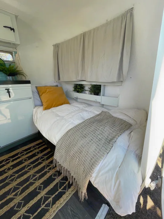 Cozy RV rental bedroom features a white bed with a yellow pillow, a textured blanket, and built-in shelves with plants.. Boler Holitrai 1976