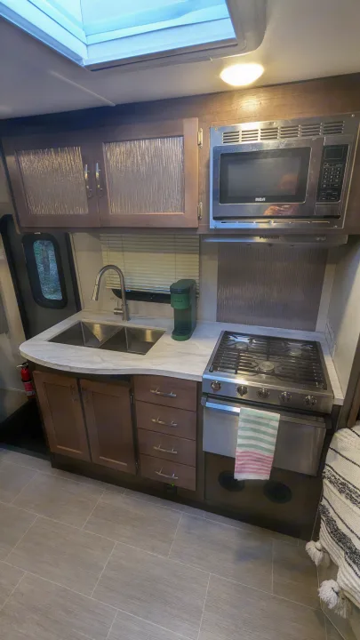 Functional RV rental kitchen with stainless sink, RCA microwave, Dometic stove, Keurig, wood-look floor, and storage.. Adventurer Ford Transit 2020