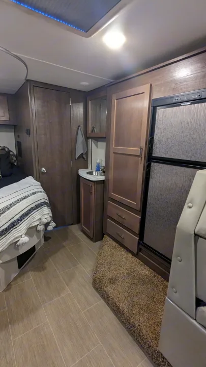 Modern RV rental interior: bedroom area, mini-sink, fridge, cabinets, and distinct tile and carpeted flooring.. Adventurer Ford Transit 2020