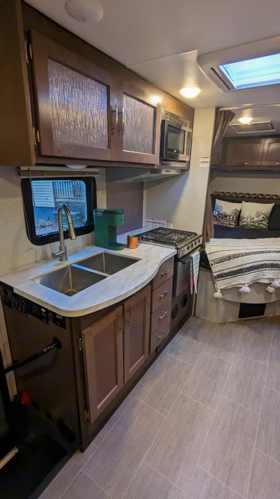 This rv rental interior features a compact kitchen with a sink, stove, microwave, & coffee maker, plus a cozy bed area.. Adventurer Ford Transit 2020