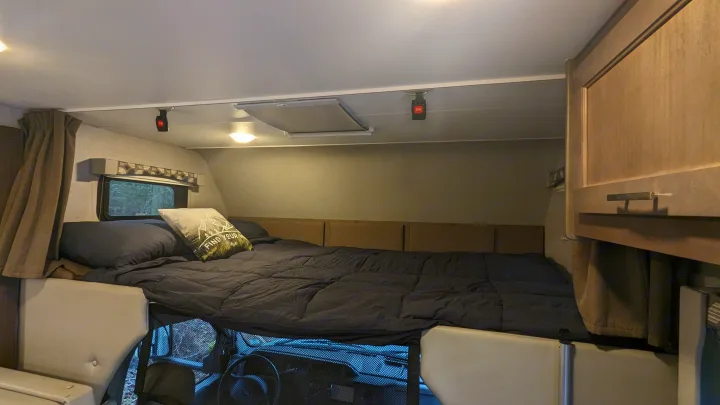 Interior of an RV rental: a large cab-over bed with dark bedding, showing the mesh-covered windshield below.. Adventurer Ford Transit 2020