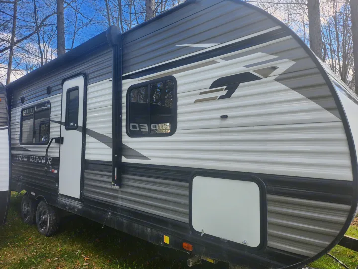 Side view of a grey and white Trail Runner travel trailer, an ideal RV rental for outdoor trips, parked under trees.. Heartland RVs Trail Runner 2022
