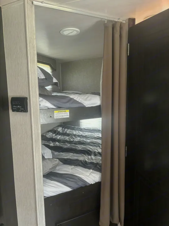 Inside an RV rental, showing stacked bunk beds with striped bedding, a privacy curtain, ceiling vent, and thermostat.. Heartland RVs North Trail 2018